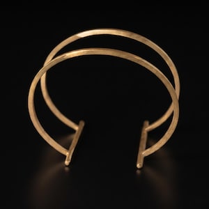 May include: A gold-toned cuff bracelet featuring two parallel curved bands that connect to two vertical bars at the open ends. The metal surface has a subtle hammered texture, creating a reflective finish on the circular structure of the jewelry.