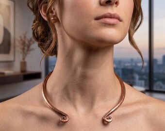 May include: A hammered copper torque necklace featuring a rigid, open-ended circular band with decorative spiral terminals at each end. The metal surface displays a textured, rustic finish with warm reddish-brown tones that rest around the neck.
