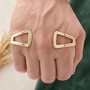 Può includere: Un bracciale rigido in metallo color oro caratterizzato da due estremità aperte di forma geometrica trapezoidale. La superficie lucida e liscia del bracciale riflette la luce, creando un aspetto metallico brillante su tutta la struttura curva.
