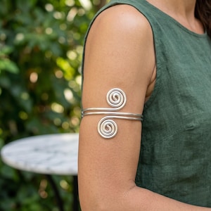 May include: A silver-colored metal arm cuff featuring a double-wire design that wraps around the upper arm. The ends of the wire are shaped into two distinct, flat circular spirals that face in opposite directions, creating a symmetrical look.