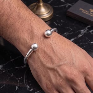 May include: A polished silver cuff bracelet featuring a smooth, open-ended design with two large, spherical ball terminals at each end. The bracelet has a reflective, metallic finish and a simple, circular band structure that rests on the wrist.