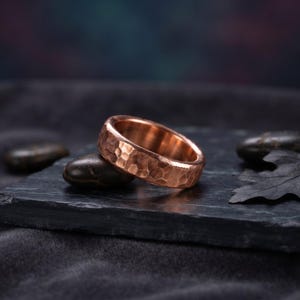 Könnte beinhalten: Ein Kupferring mit einer gehämmerten, texturierten Oberfläche. Der Ring hat ein abgerundetes Profil mit einem metallischen, rotbraunen Finish. Der Ring liegt auf einer dunklen, flachen Oberfläche und zeigt die reflektierende Qualität des Metalls sowie die durch den Hammervorgang entstandenen Vertiefungen auf der Außenseite des Bandes.