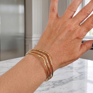 Puede incluir: Un conjunto de tres brazaletes de metal en tono dorado con un diseño en forma de V. Cada brazalete presenta una superficie texturizada y martillada que refleja la luz, creando una apariencia superpuesta al llevarlos juntos en la muñeca.