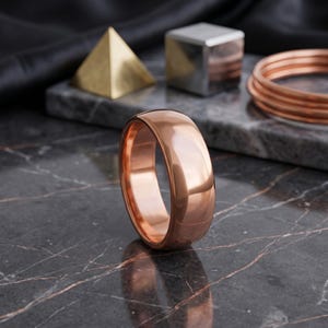 Puede incluir: Un anillo de banda en color oro rosa pulido con una superficie lisa y abovedada. El anillo presenta un acabado metálico reflectante y un perfil redondeado, colocado verticalmente sobre una superficie de mármol oscuro veteado con brillo.