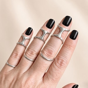 May include: A collection of silver-toned rings worn on the fingers. Three of the rings feature a distinctive H-shaped design that spans the middle joint, while two additional plain, smooth silver bands are worn on the lower part of the fingers.