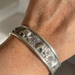 May include: Silver cuff bracelet featuring embossed elephant designs and floral patterns. The bracelet has a slightly tarnished finish, giving it a vintage appearance. The cuff is approximately 2.5 inches wide.