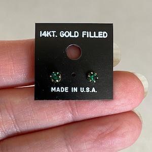 May include: A pair of small stud earrings with round, emerald-green gemstones set in gold-filled settings. The earrings are displayed on a black card that reads "14kt. Gold Filled" and "Made in U.S.A."