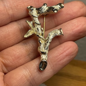 May include: A gold-toned metal brooch shaped like a textured bamboo branch with small leaves. The surface of the metal features a detailed, realistic bark-like texture with distinct nodes and cut ends, and a thin metal pin is visible on the back.