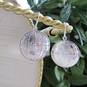 May include: A pair of silver-toned globe earrings featuring a detailed map of the world etched onto the surface of each spherical charm. The earrings are suspended from silver-toned metal hooks, showcasing a spherical design with longitude and latitude lines.