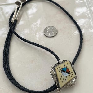 May include: A bolo tie featuring a black braided leather cord with silver tips and a diamond-shaped silver pendant. The pendant contains an inlaid sunburst design with mother-of-pearl, turquoise, red coral, and black stone, shown next to a US quarter.