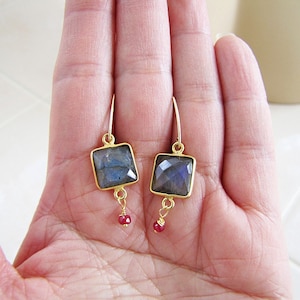 May include: A pair of gold-toned earrings. Each earring features a square, faceted, gray-blue stone set in a gold frame. A small, red, round stone dangles below each square stone. The earrings are displayed on a light-colored background.