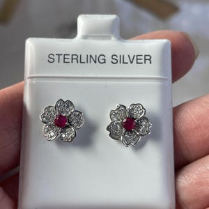 May include: A pair of sterling silver stud earrings shaped like five-petaled flowers. Each earring features a central round red gemstone surrounded by small clear stones set into the silver petals. The earrings are mounted on a white display card.