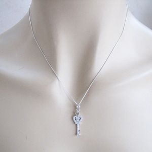 May include: A silver-toned necklace featuring a delicate chain and a small, key-shaped pendant. The pendant is encrusted with tiny, clear, sparkling stones along its bow and shaft, creating a bright, reflective surface against the metal finish.