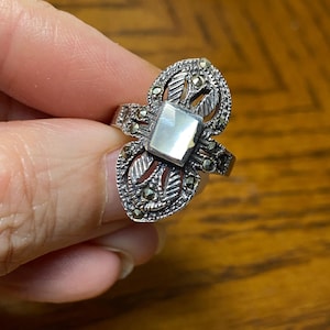 May include: A silver ring with a square mother of pearl stone surrounded by marcasite stones. The ring has a delicate, intricate design.