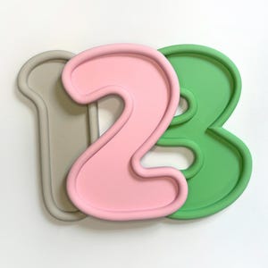 Puede incluir: Un conjunto de tres cortadores de galletas con forma de números coloridos. Los números están en un estilo de fuente de burbujas. Los números son de color gris claro, rosa y verde. Los números están dispuestos en el orden 1, 2 y 3.