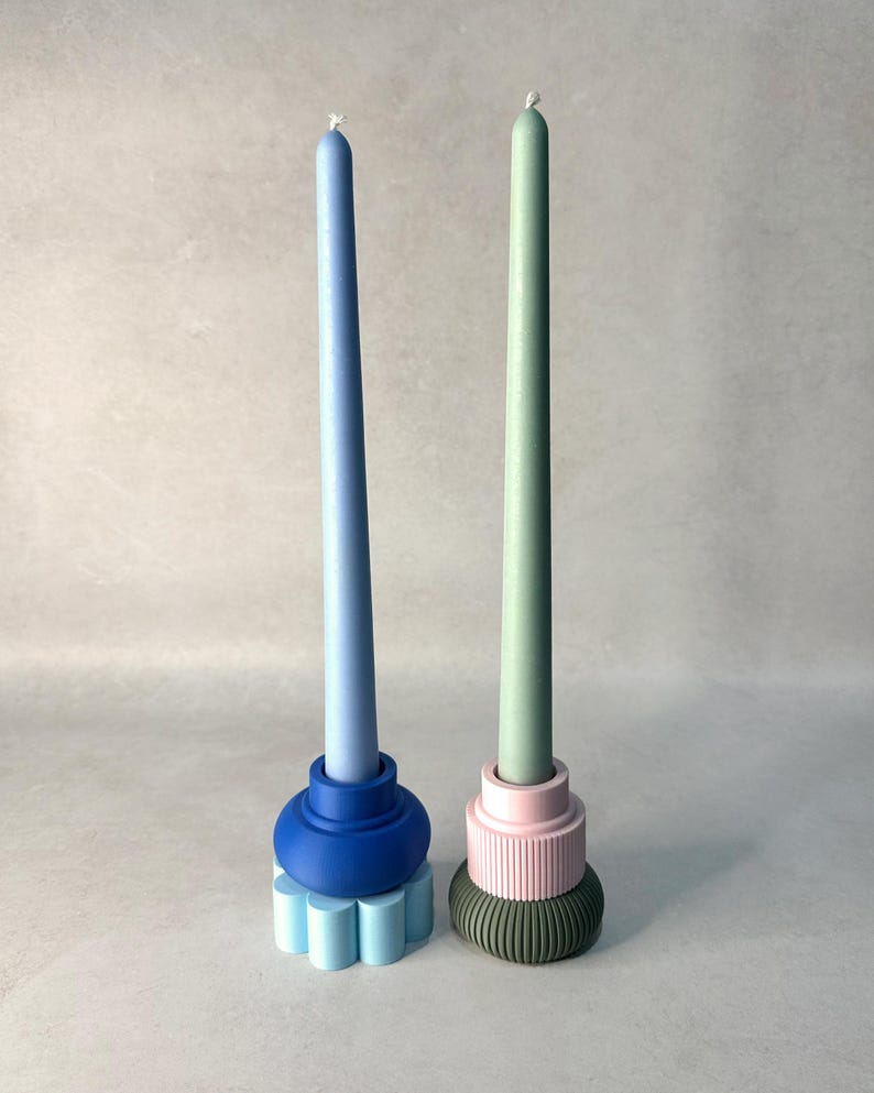 May include: Two decorative candles. One is light blue, atop a blue base with a flower-like design. The other is green, on a pink and green base. The candles are slender and stand on a light gray surface.