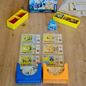 Op de afbeelding: Bordspel "Castle Combo" met een blauwe doos met kasteelillustraties. Het spel bevat kaarten, fiches en gele en blauwe opbergbakken. Het spel staat op een houten oppervlak.