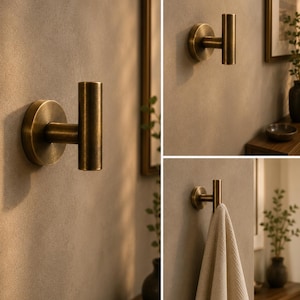 May include: A wall-mounted hook with a cylindrical design and a brushed brass finish. The hook features a circular base plate and a vertical post, providing a functional point for hanging items. The metal surface has a warm, muted golden tone.