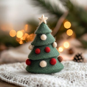 Op de afbeelding: Een handgemaakte, groene vilten kerstboom met een ster erbovenop. De boom is versierd met rode, witte en bruine pompons. Hij staat op een wit, getextureerd oppervlak, met vage lichten op de achtergrond, wat een feestelijke scène creëert.