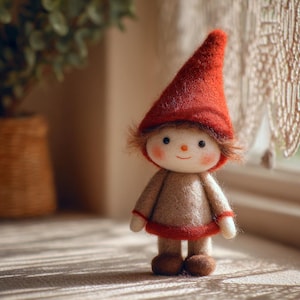 May include: A handmade felt gnome doll with a red pointed hat, brown hair, and a smiling face. The doll wears a tan outfit with red trim. The doll is standing on a light-colored surface near a window.