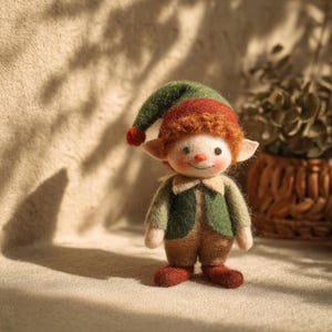 May include: A handcrafted felt elf doll with a green hat, red hair, and a green vest over a brown outfit. The doll has a smiling face with a red nose and rosy cheeks. The elf is standing on a neutral surface.