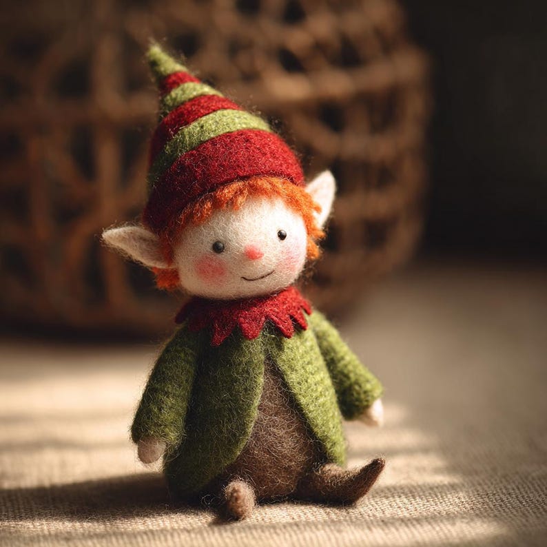 May include: A handmade felt elf doll with a red and green striped hat, green jacket, and brown pants. The doll has white ears, orange hair, and a smiling face with rosy cheeks. The elf is sitting on a neutral-colored surface.
