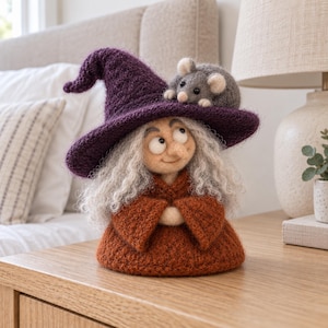 Könnte beinhalten: Eine gefilzte Figur, die einen Charakter mit einem dunkelvioletten spitzen Hut und einem rostfarbenen Gewand darstellt. Die Figur hat lockiges graues Haar, eine kleine Nase und große Augen. Eine kleine graue Maus sitzt oben auf dem violetten Hut.