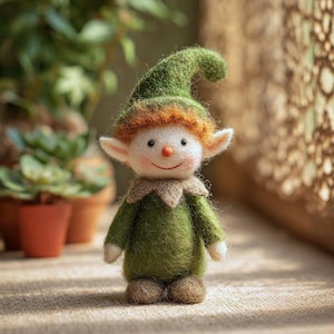 May include: A handcrafted felt elf doll with a green hat and outfit, red hair, and a smiling face. The doll has white ears, a tan collar, and brown feet. It is standing on a textured surface with plants in the background.