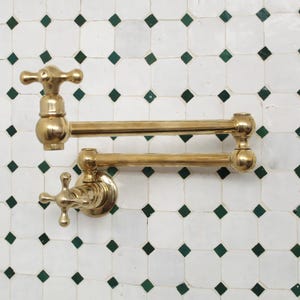 Op de afbeelding: Een messing potvuller kraan met een arm met twee gewrichten is gemonteerd op een witte tegelwand met groene diamantaccenten. De kraan heeft kruisvormige handgrepen en een gepolijste afwerking.