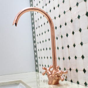 Può includere: Un rubinetto color rame con un beccuccio curvo e maniglie ornate. Il rubinetto è montato su un piano di lavoro colorato chiaro, con un lavandino bianco sottostante. Lo sfondo presenta piastrelle bianche con dettagli a diamante verdi.