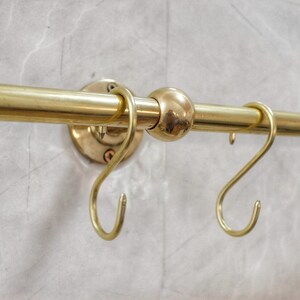 May include: A brass-colored kitchen rail with several S-shaped hooks. The rail is mounted on a light gray, textured wall. The rail has a round ball in the middle and is attached to the wall with round mounting plates.