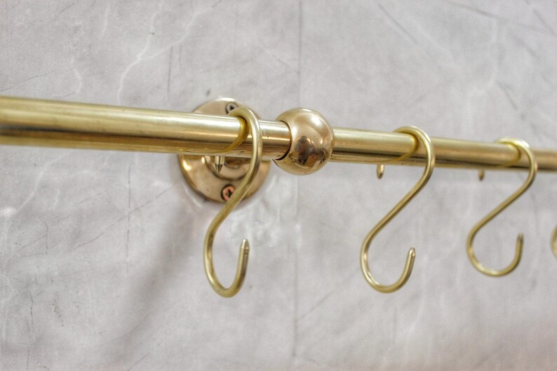 May include: A brass-colored kitchen rail with several S-shaped hooks. The rail is mounted on a light gray, textured wall. The rail has a round ball in the middle and is attached to the wall with round mounting plates.