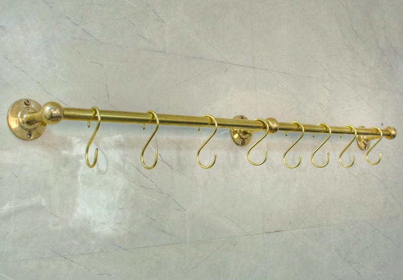 May include: A brass-colored, wall-mounted rack with eight curved hooks. The rack has a horizontal bar with decorative end caps and mounting hardware. The hooks are evenly spaced along the bar, suitable for hanging items. The rack is mounted on a light-colored wall.