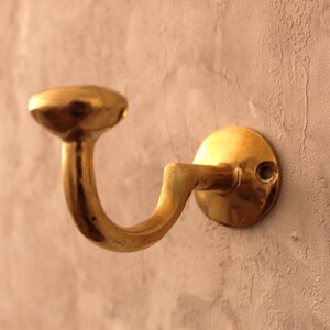 May include: A brass-colored wall hook with a curved design. The hook has a circular base and a rounded top, mounted on a textured, light-colored wall. The hook is designed to hold items and adds a touch of vintage style.