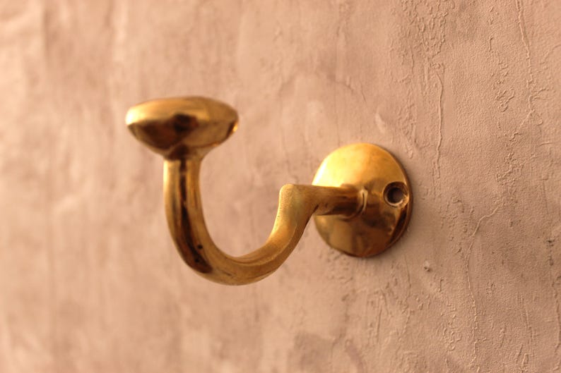 May include: A brass-colored wall hook with a curved design. The hook has a circular base and a rounded top, mounted on a textured, light-colored wall. The hook is designed to hold items and adds a touch of vintage style.