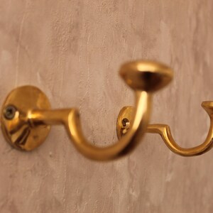 May include: A gold-colored, double-hook wall-mounted coat rack. The rack has a curved design with two hooks and a circular base. The finish is shiny and reflective, and the rack is mounted on a textured wall.