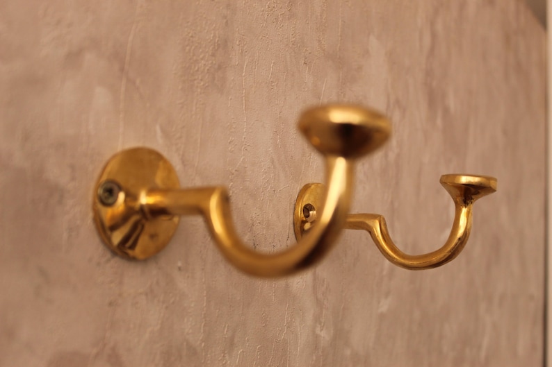 May include: A gold-colored, double-hook wall-mounted coat rack. The rack has a curved design with two hooks and a circular base. The finish is shiny and reflective, and the rack is mounted on a textured wall.