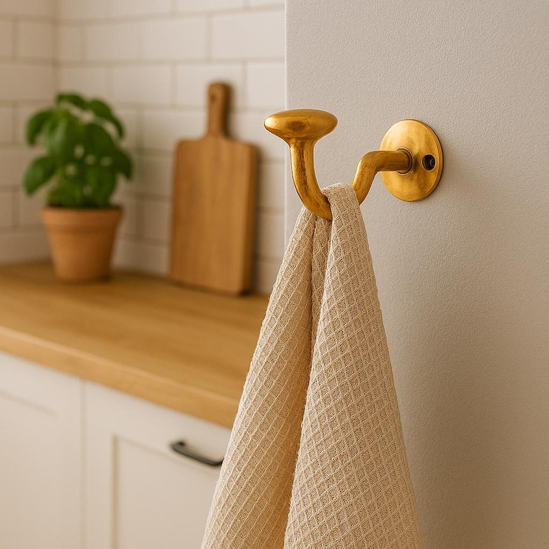 May include: A gold-colored, curved hook mounted on a wall, holding a beige waffle-weave towel. The hook has a circular base and a rounded top. The background includes a wooden cutting board and a potted plant.