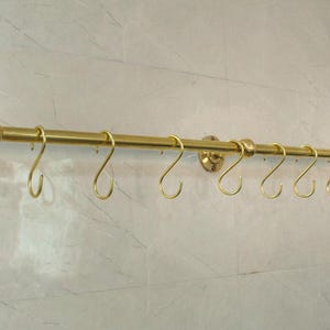 May include: A brass-colored kitchen rail with eight S-shaped hooks. The rail is mounted on a wall and has decorative end caps and mounting plates. The hooks are evenly spaced along the rail, providing a stylish storage solution.