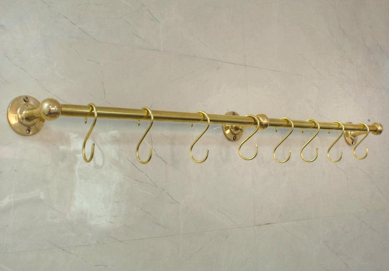 May include: A brass-colored kitchen rail with eight S-shaped hooks. The rail is mounted on a wall and has decorative end caps and mounting plates. The hooks are evenly spaced along the rail, providing a stylish storage solution.