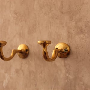 May include: Two antique brass wall hooks with a curved design. The hooks are mounted on a textured, light brown wall. The hooks have a circular base and a curved arm that ends in a small, flat disc.