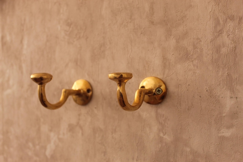 May include: Two antique brass wall hooks with a curved design. The hooks are mounted on a textured, light brown wall. The hooks have a circular base and a curved arm that ends in a small, flat disc.