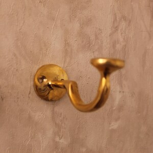 May include: A brass-colored wall-mounted handrail bracket. The bracket has a circular base, a curved arm, and a rounded top. The metal has a polished finish. The bracket is mounted on a textured, light brown wall.