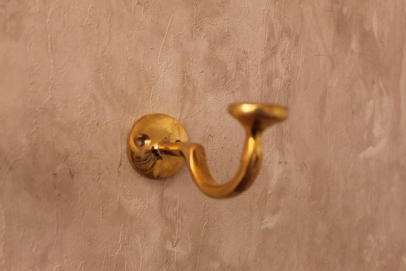 May include: A brass-colored wall-mounted handrail bracket. The bracket has a circular base, a curved arm, and a rounded top. The metal has a polished finish. The bracket is mounted on a textured, light brown wall.