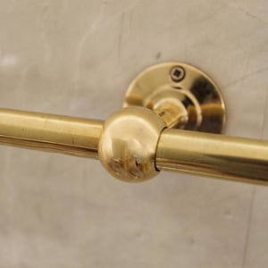 May include: A close-up of a gold-colored towel bar. The bar is cylindrical with a rounded center and is attached to the wall with a circular mounting plate. The finish is shiny and reflective.