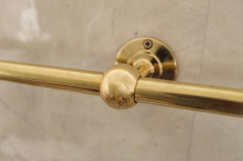 May include: A close-up of a gold-colored towel bar. The bar is cylindrical with a rounded center and is attached to the wall with a circular mounting plate. The finish is shiny and reflective.