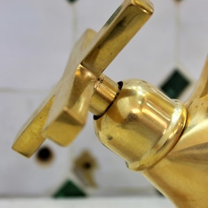 May include: Close-up of a gold-coloured tap with a cross-shaped handle. The tap has a shiny, reflective surface and is set against a backdrop of white tiles with green and black diamond-shaped accents. The tap is a bathroom fixture.