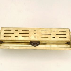 May include: A rectangular brass box with a hinged lid. The lid features a series of horizontal and circular cutouts. The box has a simple, utilitarian design with a tarnished finish, suggesting age or use. The box is likely a container or storage item.