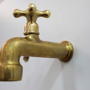 May include: A brass faucet with a cross-shaped handle is mounted on a white wall. The faucet has a curved spout and a circular base. The brass has a warm, golden hue.