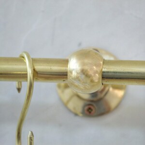 May include: Close-up of a brass-colored curtain rod with a rounded finial and a hook. The rod is mounted on a circular base with a screw. The metal has a polished finish, reflecting light. The design is simple and functional.